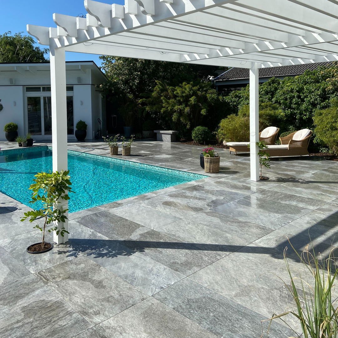 Pooldäck med grå klinker, vit pergola och grönska kring uteplatsen.